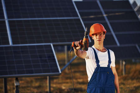 Cordless screwdriver in hand. Male worker in blue uniform outdoors with solar batteries at sunny day.の写真素材