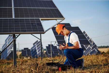 Using special equipment. Male worker in blue uniform outdoors with solar batteries at sunny day.の写真素材
