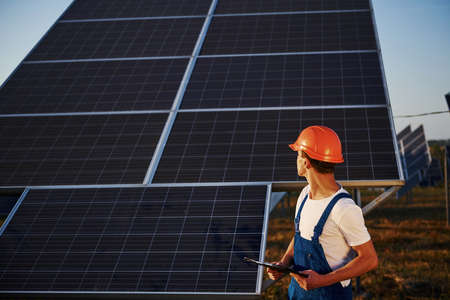 Holds notepad in hand. Male worker in blue uniform outdoors with solar batteries at sunny day.の写真素材