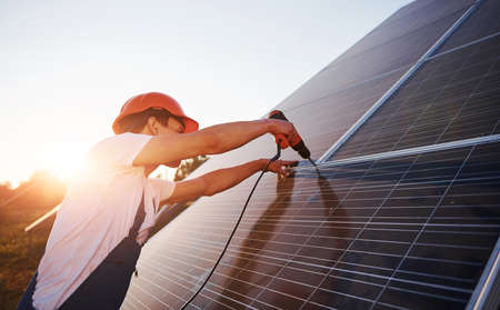 Using cordless screwdriver. Male worker in blue uniform outdoors with solar batteries at sunny day.の写真素材