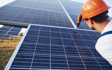 Using cordless screwdriver. Male worker in blue uniform outdoors with solar batteries at sunny day.