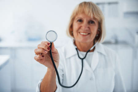 Holds stethoscope in hand. Portrait of senior doctor in white uniform that stands in the clinic.の写真素材