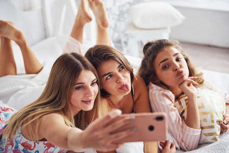 Making selfie by using pink phone. Happy female friends having good time at pajama party in the bedroom.の写真素材