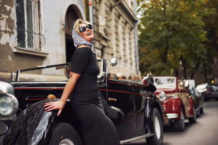 Blonde woman in sunglasses and in black dress leaning on old vintage classic car.の写真素材
