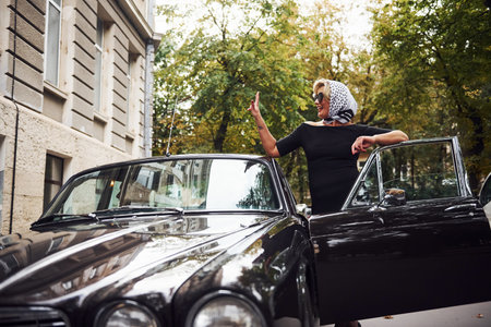 Blonde woman in sunglasses and in black dress near old vintage classic car.