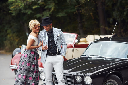 Beautiful couple in old fashioned wear near retro car with forest at background.