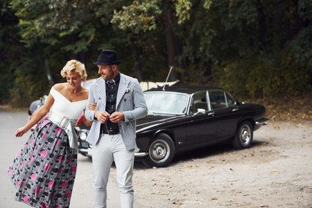 Beautiful couple in old fashioned wear near retro car with forest at background.