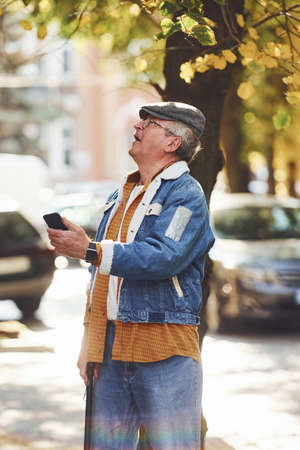 Stylish senior in fashionable clothes and in glasses stands with phone in the city.の写真素材