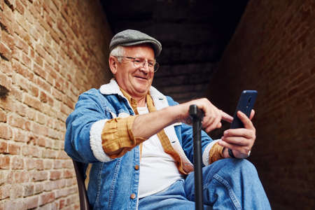 Stylish senior in fashionable clothes and in glasses sits on the chair and uses phone.の写真素材