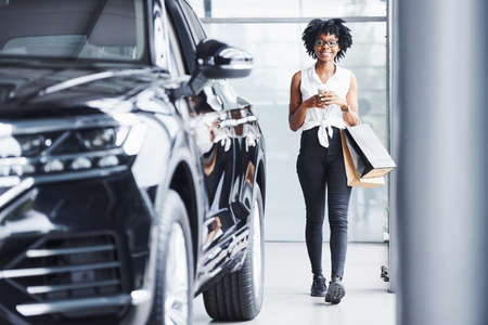 Young african american woman in glasses stands in car salon near vehicle with package in hands.の写真素材