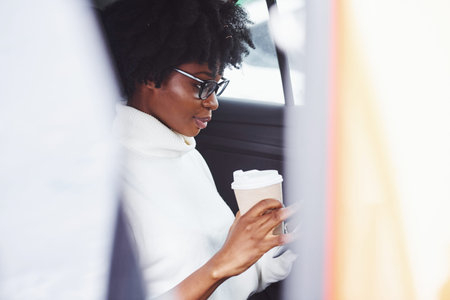 With cup of drink. Young african american woman sits inside of new modern car.