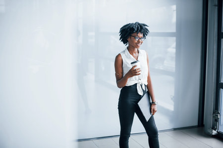 Young african american woman in glasses stands indoors in the office with tablet in hands.