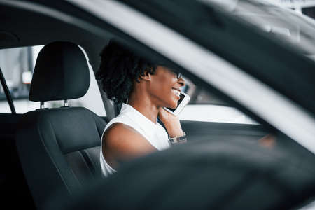 Using phone. Young african american woman sits inside of new modern car.の写真素材
