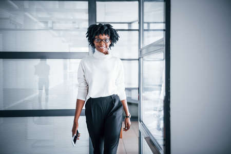 Young african american woman in glasses stands indoors in the office with phone.の写真素材