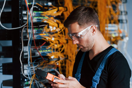Young man in protective glasses works with internet equipment and wires in server room.