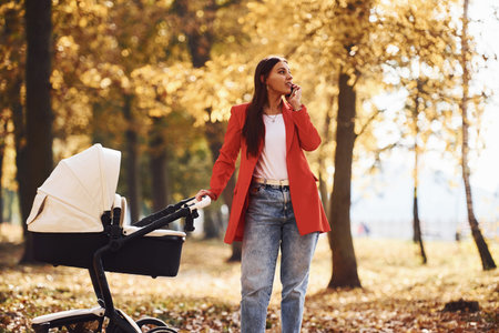 Talking by the phone. Mother in red coat have a walk with her kid in the pram in the park at autumn...