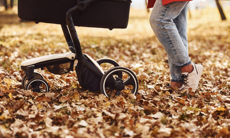 Close up view. Mother in red coat have a walk with her kid in the pram in the park at autumn time.