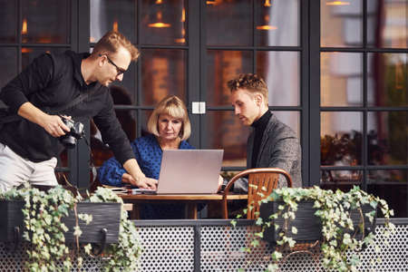 Photographer with camera talks to people. Young guy in formal clothes have a business talk with old woman in cafe.の写真素材