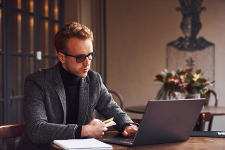 Elegant young guy in formal wear sits in cafe with his laptop and credit card in hand.