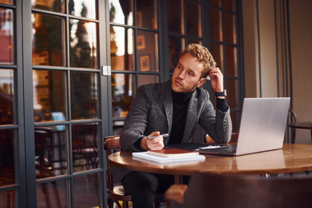 Elegant young guy in formal wear sits in cafe with his laptop.