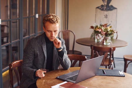Elegant young guy in formal wear sits in cafe with his laptop and talk by the phone.の写真素材
