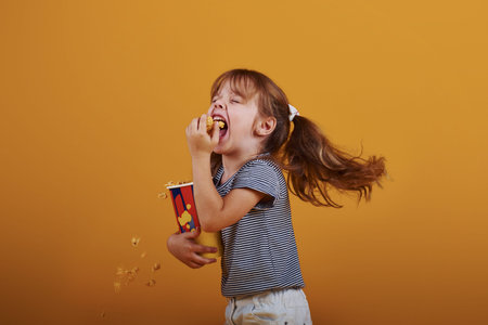 Cute little girl in in the studio eats popcorn and stands against yellow background.の写真素材