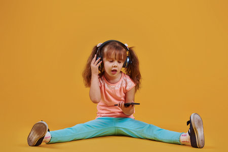Portrait of cute little girl in headphones and with phone in the studio against yellow background.