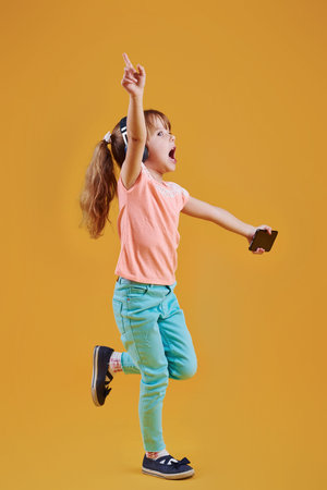 Portrait of cute little girl in headphones and with phone in the studio against yellow background.の写真素材