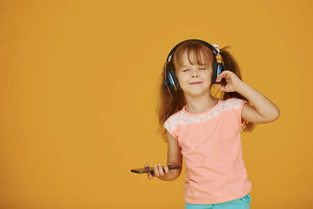 Portrait of cute little girl in headphones and with phone in the studio against yellow background.の写真素材