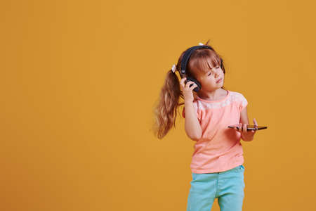 Portrait of cute little girl in headphones and with phone in the studio against yellow background.の写真素材