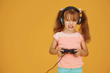 Little female gamer in headphones and with joystick in hands playing video games against yellow background.の写真素材