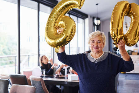 With balloons of number 60 in hands. Senior woman with family and friends celebrating a birthday indoors.の写真素材