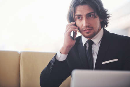 Portrait of handsome young businessman in black suit and tie sits and have a call.の写真素材