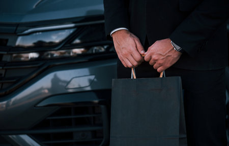 Close up view of businessman in black formal wear near car and with shopping bags in hands.