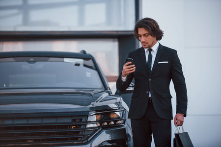Portrait of handsome young businessman in black suit and tie outdoors near modern car and with...