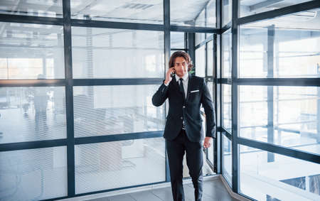 Talking by the phone in the office. Portrait of handsome young businessman in black suit and tie.の写真素材