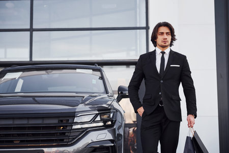 Portrait of handsome young businessman in black suit and tie outdoors near modern car and with...