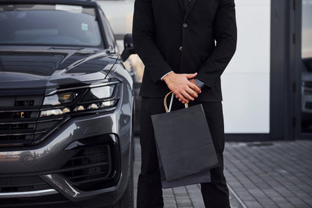 Close up view of businessman in black suit and tie stands near modern automobile with shopping bags...