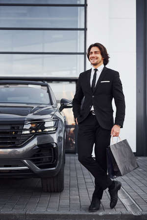 Portrait of handsome young businessman in black suit and tie outdoors near modern car and with shopping bags.の写真素材
