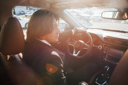 Sunny weather. Young businessman in black suit and tie driving modern automobile.