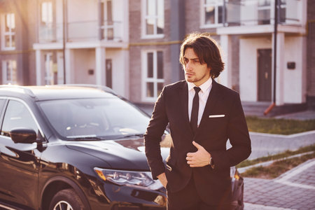 Portrait of handsome young businessman in black suit and tie outdoors near modern car in the city.