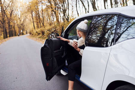 With cup of drink in hands. Girl have autumn trip by car. Modern brand new automobile in the forest.の写真素材