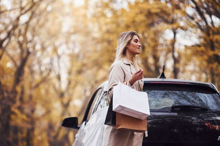 Girl walks near the car with shopping bags in hands. Modern brand new automobile in the forest.の写真素材