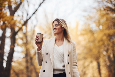 Holds cup of drink. Portrait of young brunette that is in autumn forest at daytime.の写真素材