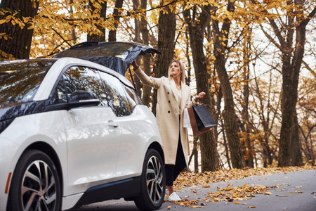 Girl with shopping bags in hands have autumn trip by car. Modern brand new automobile in the forest.