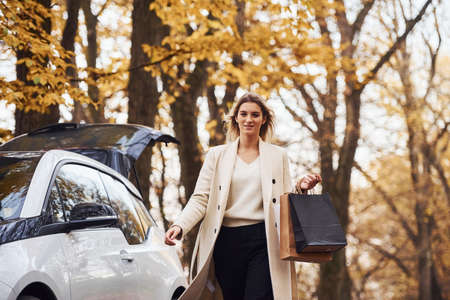 Girl walks near the car with shopping bags in hands. Modern brand new automobile in the forest.の写真素材