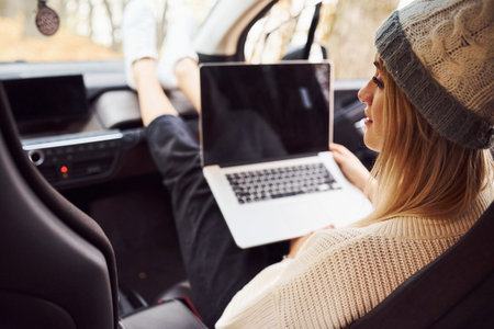 Sits with laptop and cup of drink. Girl have autumn trip by car. Modern brand new automobile in the...