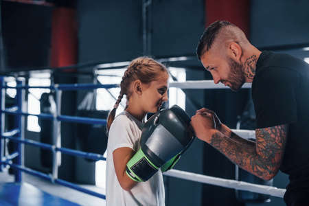 Having sparring with each other on the boxing ring. Young tattooed boxing coach teaches cute little girl in the gym.の写真素材