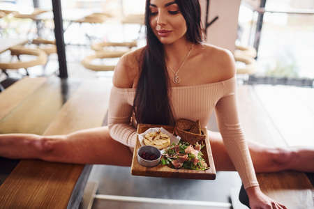 Young brunette in sexy wear soing stretches by sitting on the table and holds plate with food in restaurant.の写真素材