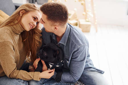 Happy young modern couple sitting on the sofa at home with christmas tree with their cute dog.の写真素材